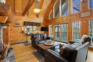 Un intérieur de chalet chaleureux avec des murs en bois, une cheminée, un coin repas et de grandes fenêtres offrant une vue sur l’extérieur.