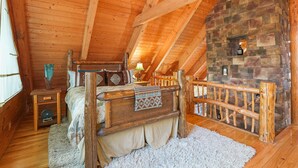 A cozy attic bedroom with a wooden ceiling, a stone fireplace, a bed with a wooden headboard, and a small table with a lamp.