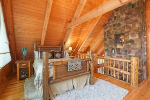 Une chambre confortable située sous les combles, avec un plafond en bois, une cheminée en pierre, un lit doté d’une tête de lit en bois et une petite table avec une lampe.