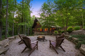 A cabin with a fire pit and two Adirondack chairs in a forest setting.