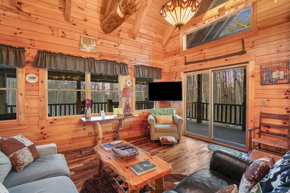 Ein gemütliches Blockhaus-Interieur mit Holzwänden, einer Couch, einem Tisch mit Büchern, einem Fernseher und einer Glastür zum Garten.