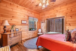 Un dormitorio de cabaña de madera con cama, cómoda, lámpara y ventana con cortinas.