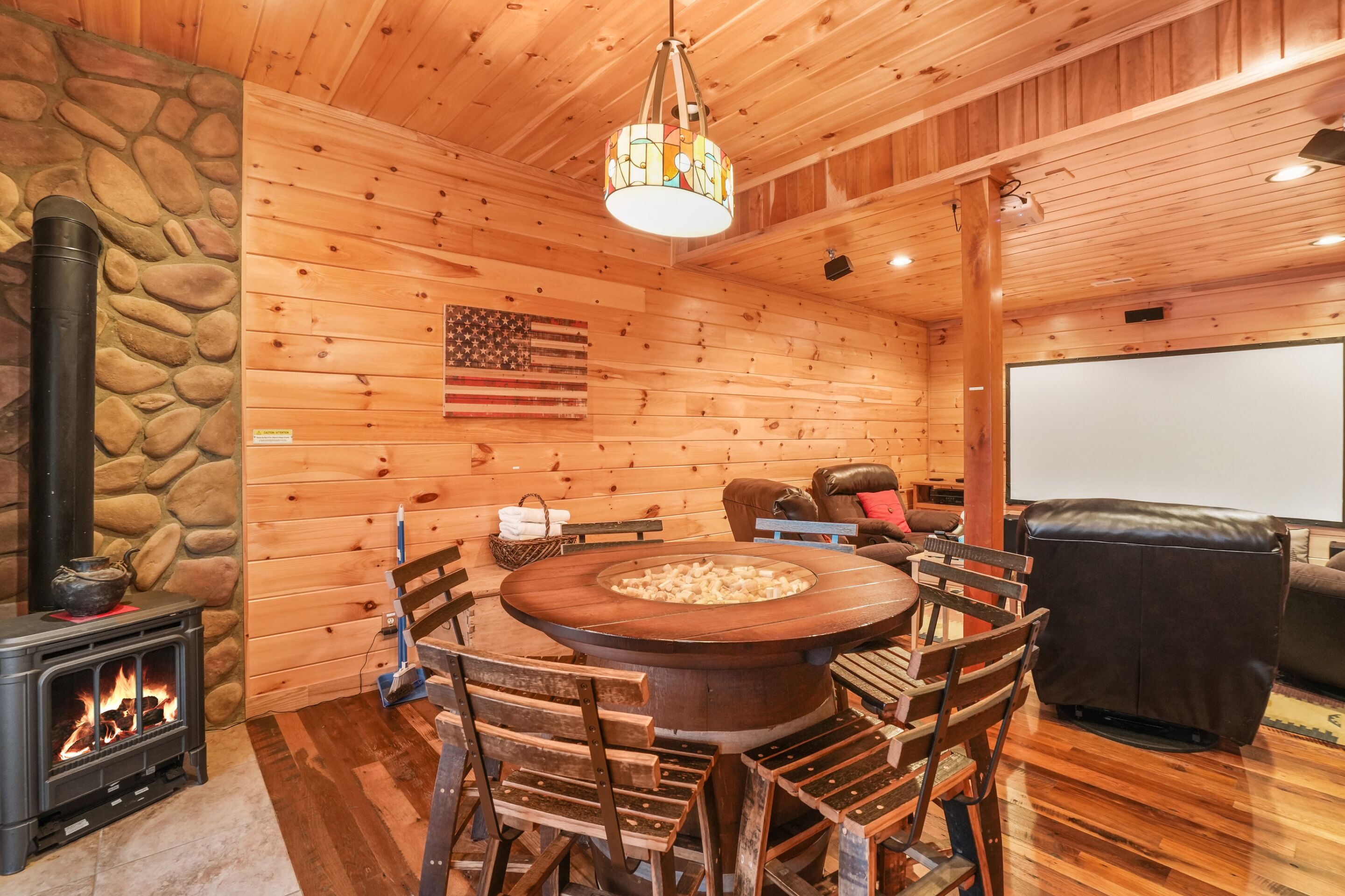 A cozy cabin interior with a stone wall, wooden walls, a fireplace, a round wooden table with chairs, a stone stove, and a large American flag.