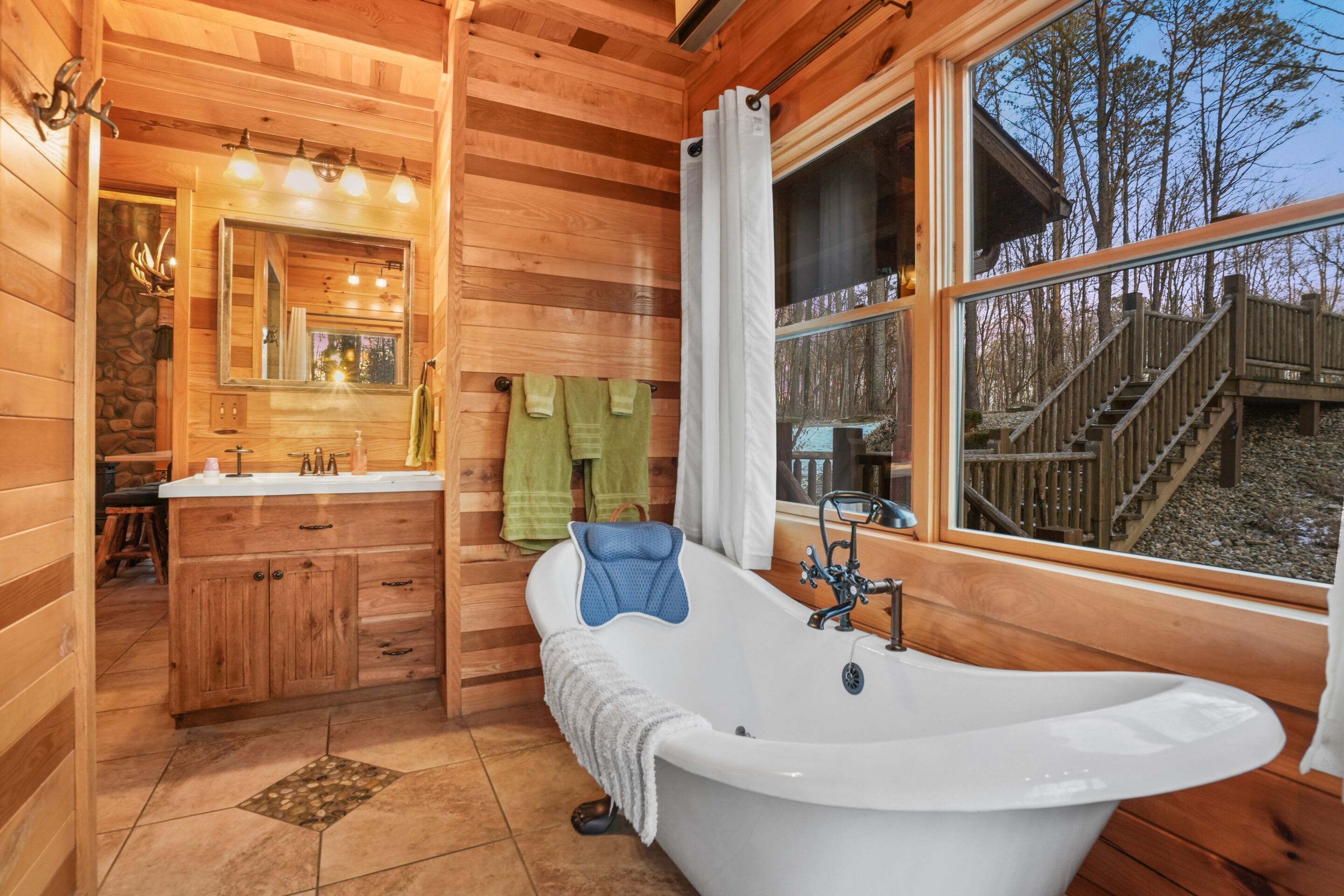Ein Badezimmer einer Holzhütte mit einer großen Badewanne, einem Spiegel und einem Fenster mit Blick auf eine Holzterrasse.
