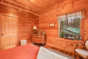 Ein gemütliches Zimmer mit Holzwänden, einem Bett mit roter Decke, einer Kommode mit Lampe und einem Fenster mit Blick auf Bäume.