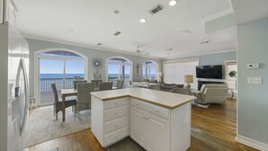 A spacious kitchen with a large island, a dining area with wicker chairs, a living room with a sofa and TV, and a balcony with a view of the ocean.
