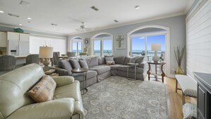 A spacious living room with a large sectional sofa, a round coffee table, and a view of the ocean through arched windows.