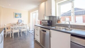 Et moderne køkken med hvide skabe, en stålvask og en opvaskemaskine. Der er et spiseområde med hvide stole og et bord, og et vindue giver udsigt til det fri.