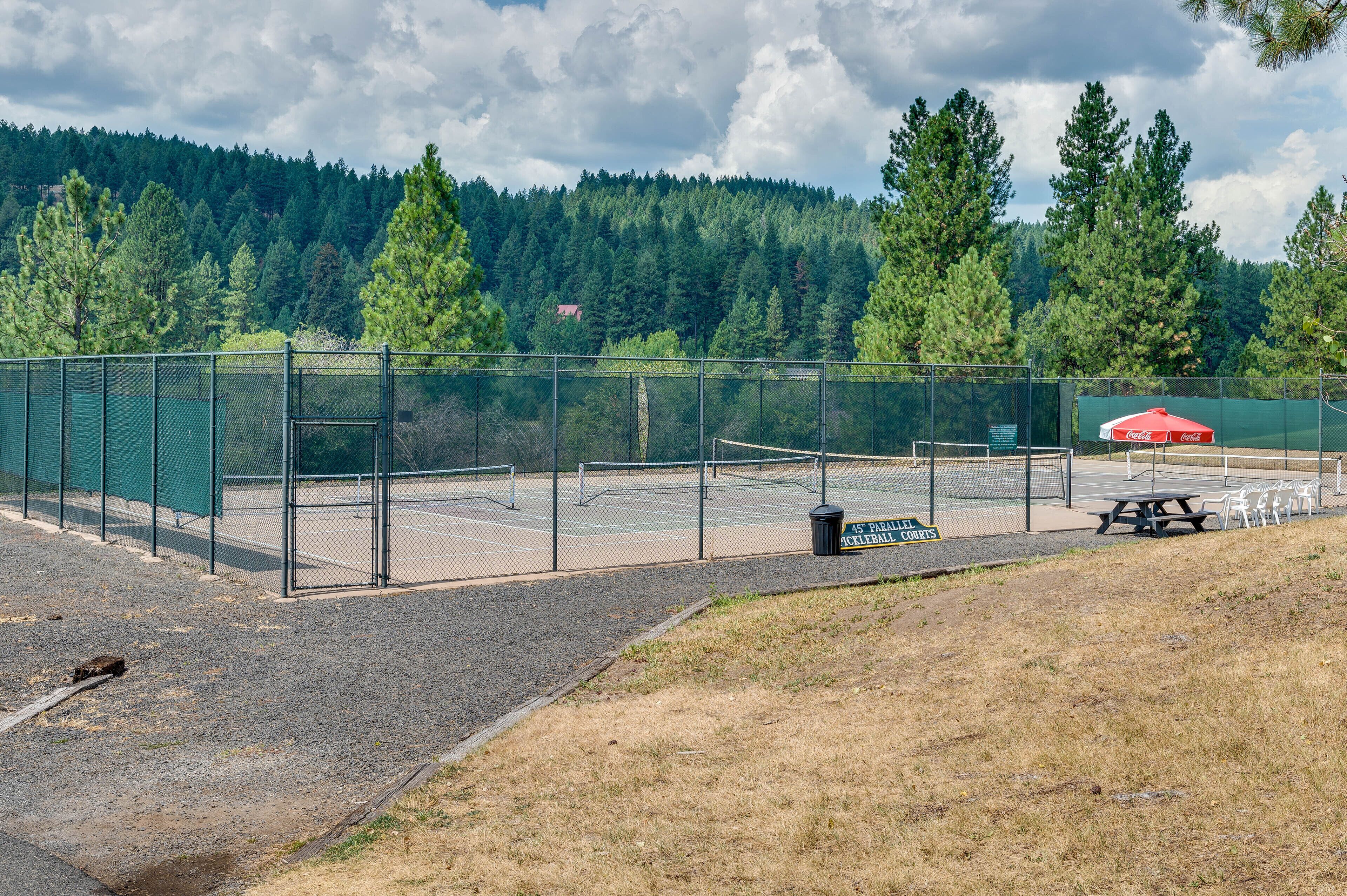 A fenced tennis court with picnic tables and a Coca-Cola branded umbrella.