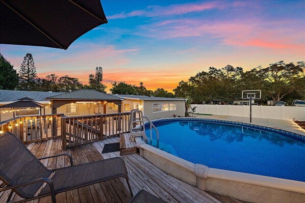 A backyard with a swimming pool, wooden deck, outdoor seating, and a basketball hoop.