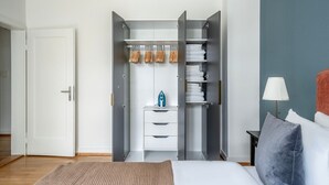 A modern bedroom with a bed, a bedside table with a lamp, a wardrobe with hanging clothes, and a small blue appliance. - Superb St. Johann 1BR w/ Storage Room, close to Station, by Blueground (Basel)