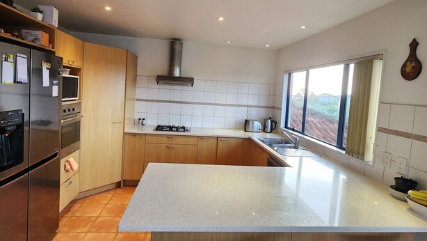 A modern kitchen with wooden cabinets, a stainless steel refrigerator, an oven, a microwave, and a gas stove. There are two bar stools and a large window providing a view of the outdoors.