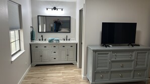 Une chambre à coucher avec un grand lit, une commode, une télévision et une salle de bain équipée d’un meuble-lavabo et d’un miroir.
