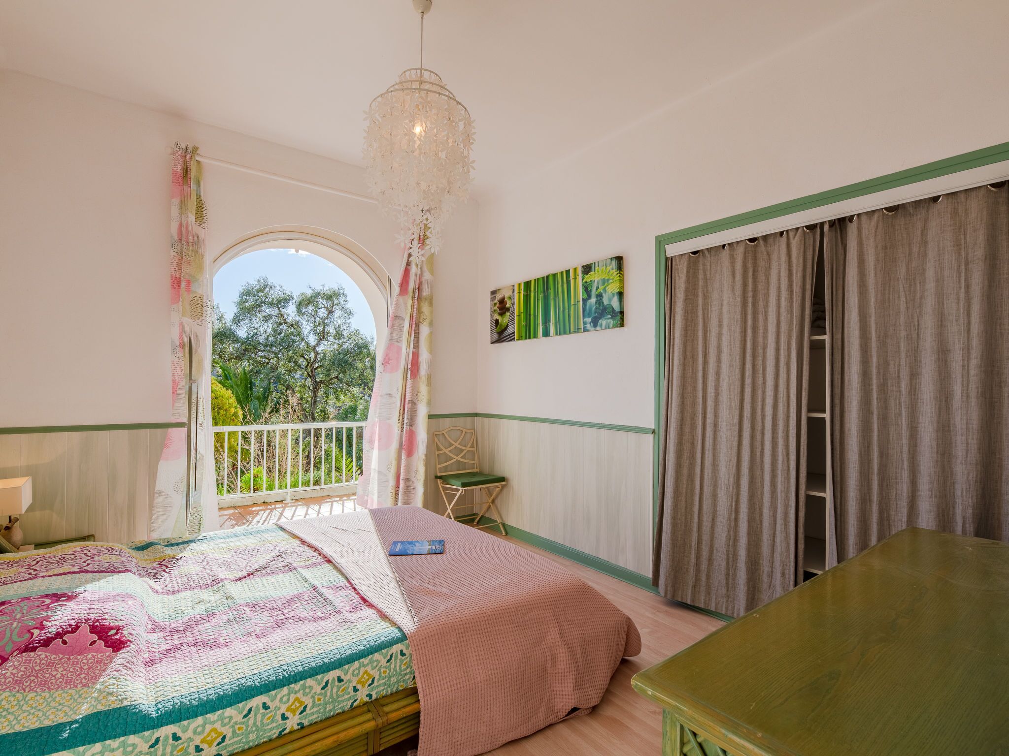 A bedroom with a bed, a green table, a window with curtains, a balcony, and a hanging light fixture.