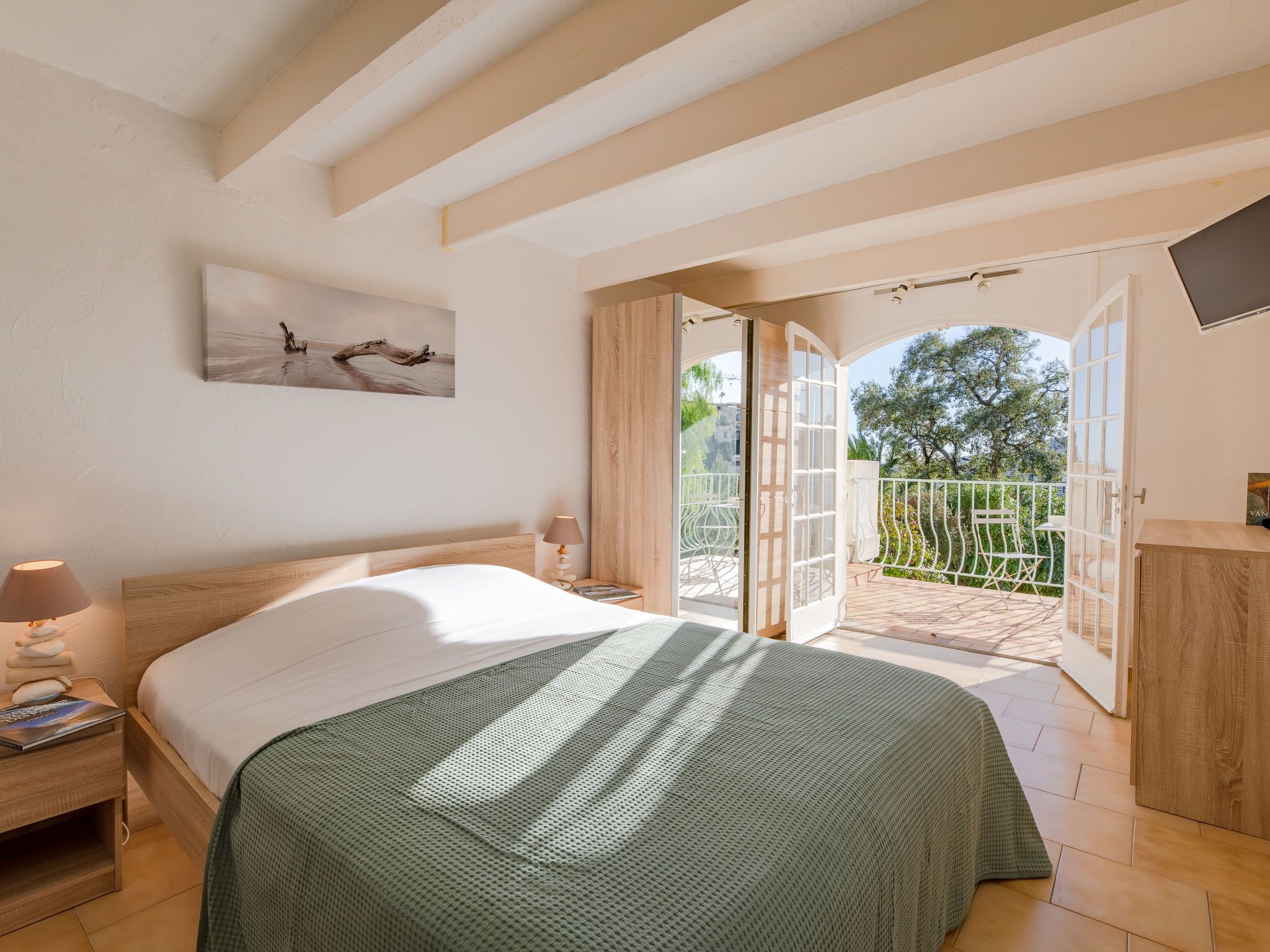 A bedroom with a bed, a nightstand, a lamp, a painting, and a balcony with a view of trees.