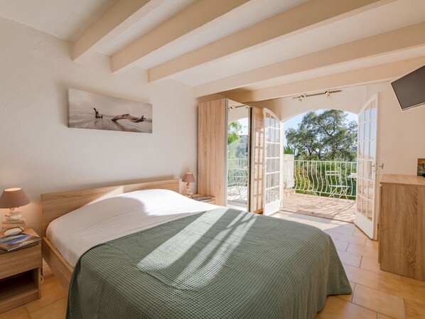 A bedroom with a bed, a nightstand, a lamp, a painting, and a balcony with a view of trees.
