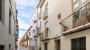 Une rue étroite, bordée de bâtiments des deux côtés, avec des balcons ornés de plantes en pot et de rampes décoratives.