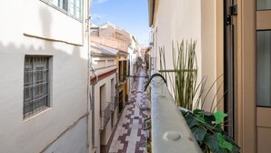 Une ruelle étroite bordée de bâtiments des deux côtés, avec un trottoir dallé et une rambarde métallique ornée de plantes.