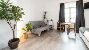 Une pièce moderne et bien éclairée, avec un canapé gris, une petite table à manger et des chaises, ainsi qu’une plante en pot.