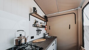 Une cuisine compacte équipée d’une cuisinière à gaz, d’une bouilloire et d’une planche à découper en bois. Des étagères au-dessus du plan de travail servent à ranger les ustensiles de cuisine, et il y a un évier avec un robinet.