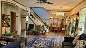 A spacious living room with a large chandelier, a dining area with wooden chairs, and a staircase with a decorative railing.