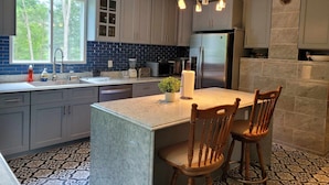 A modern kitchen with grey cabinets, a large island with two wooden chairs, a built-in oven, and a stainless steel refrigerator.