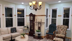A living room with a beige sofa, a round coffee table, a floral armchair, a wooden side table, a grandfather clock, and a chandelier.
