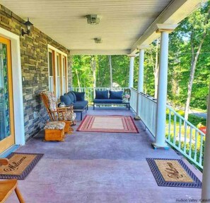 A covered porch with a stone wall, two seating areas with cushions, and a view of greenery.