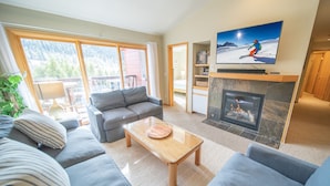 A modern living room with a fireplace, a television mounted above it, and a large window offering a view of the outdoors. - Cinnamon Ridge D323 (Keystone)