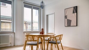 Et værelse med et rundt træbord, fire fletstole, en vase og et geometrisk abstrakt billede på væggen.