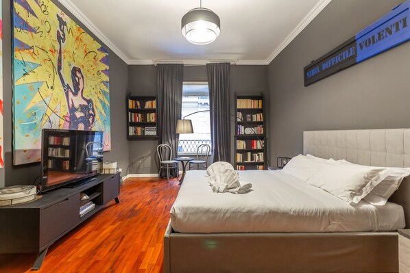 A modern bedroom with a large bed, a TV, a bookshelf, and a painting on the wall.