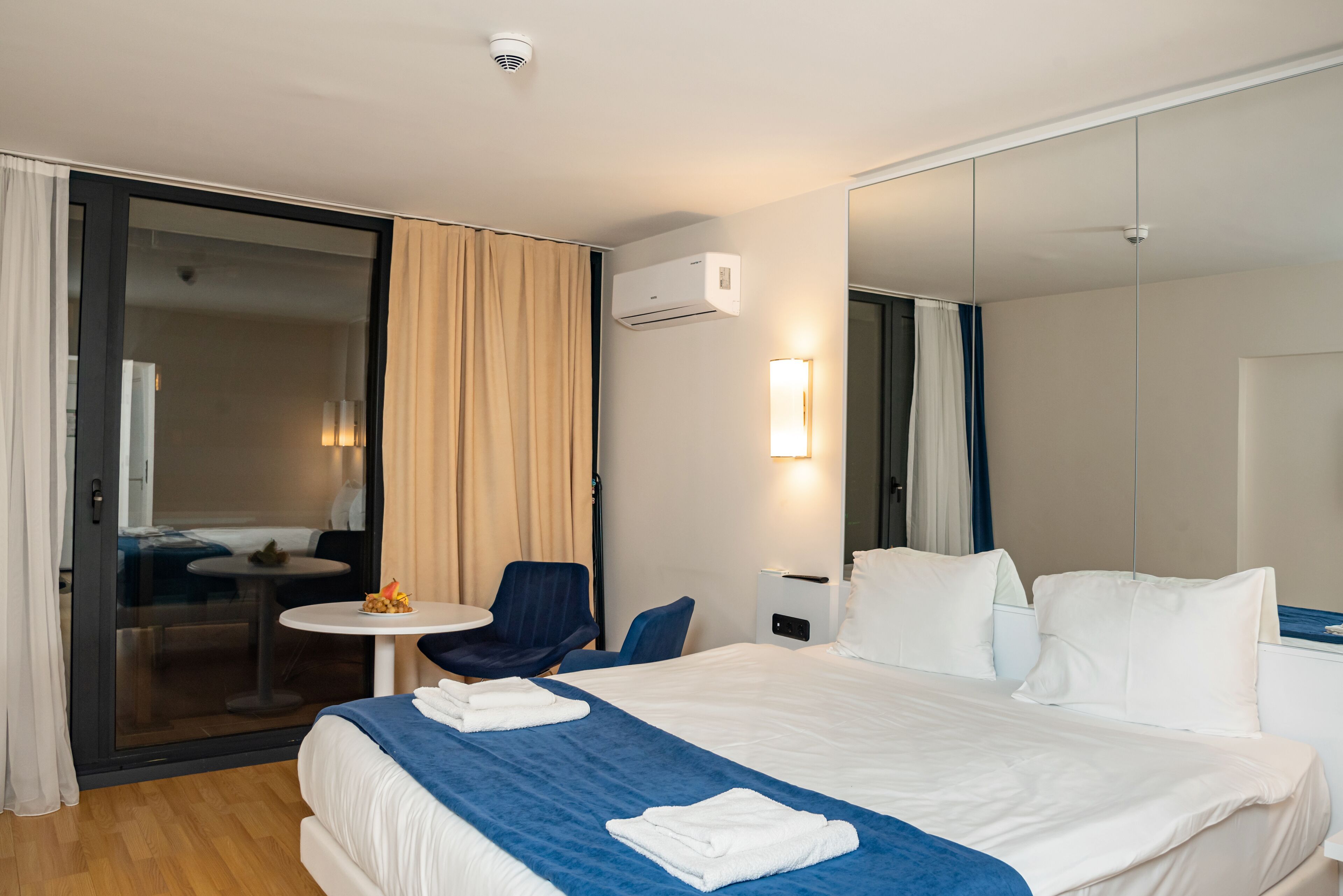 A hotel room with a bed, a small table, a chair, and a sliding glass door leading to a balcony.