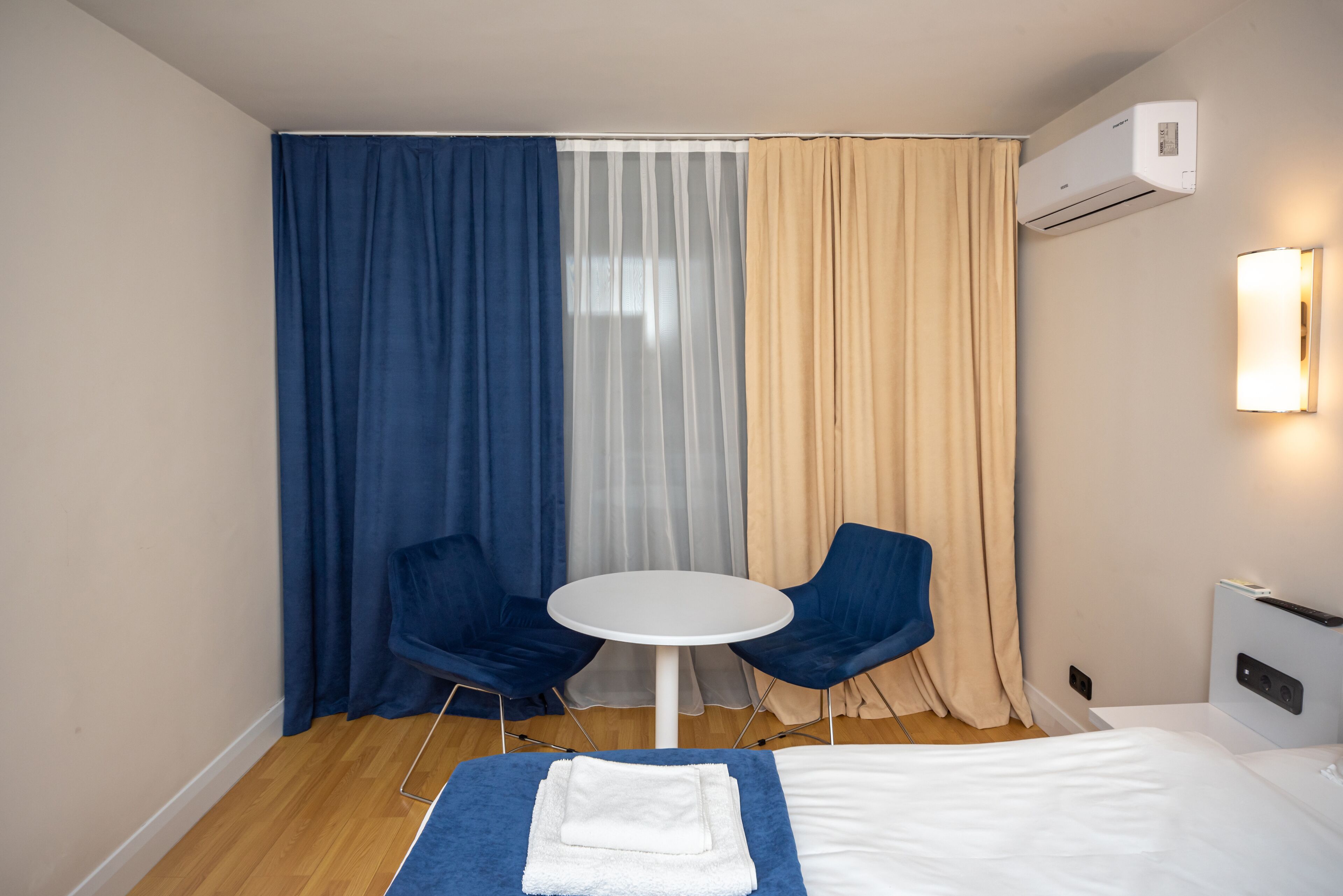 A hotel room with a bed, two blue chairs, a small round table, and a window with sheer curtains.