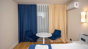 Habitación de hotel con una cama, dos sillas azules, una mesita redonda y un ventanal con cortinas traslúcidas.