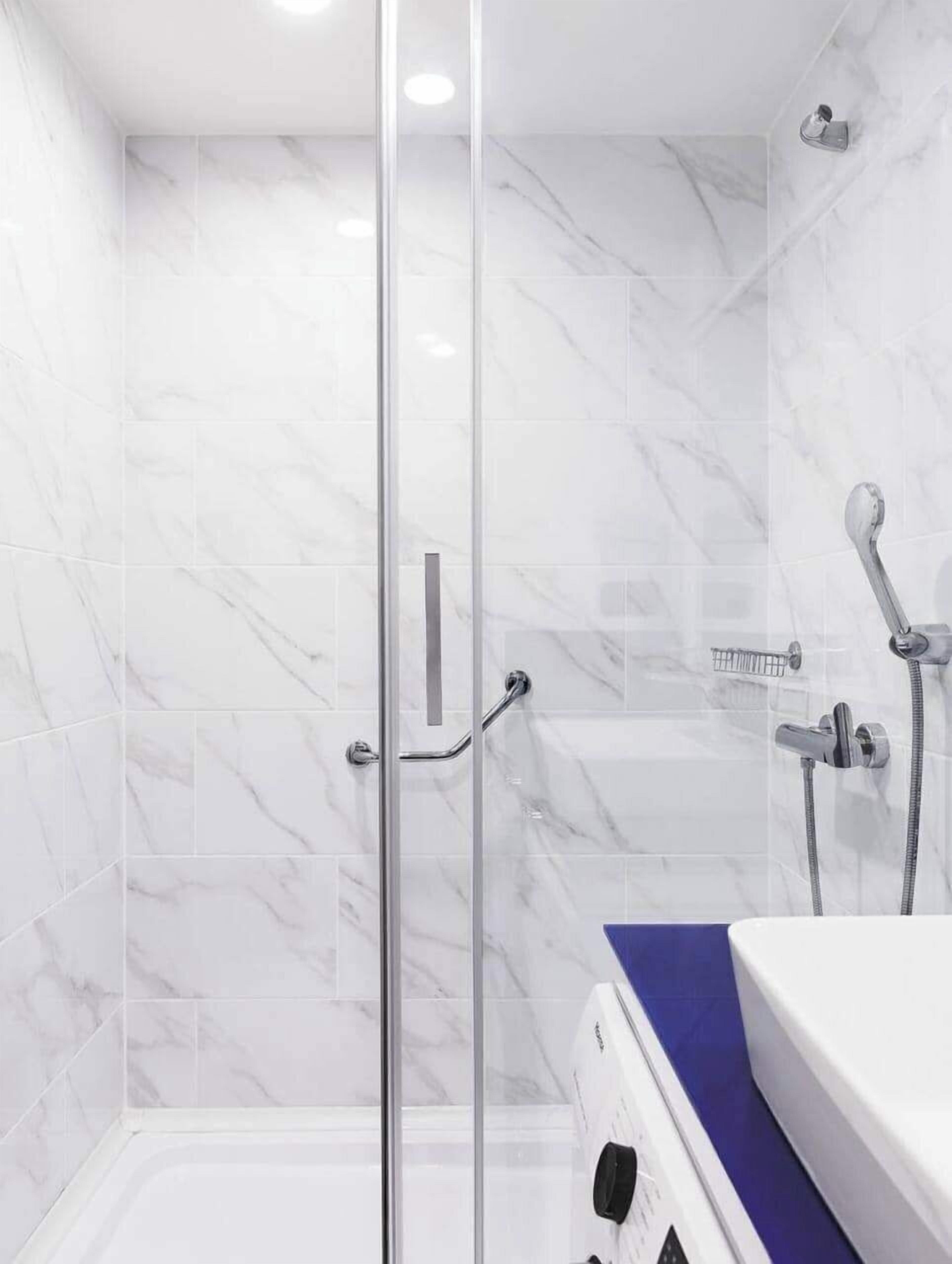 A modern bathroom with a glass shower enclosure, white marble tiles, and a blue countertop.