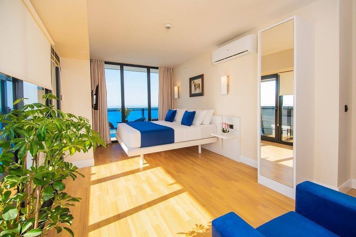 A modern hotel room with a large bed, a sofa, a potted plant, and a view of the sea.
