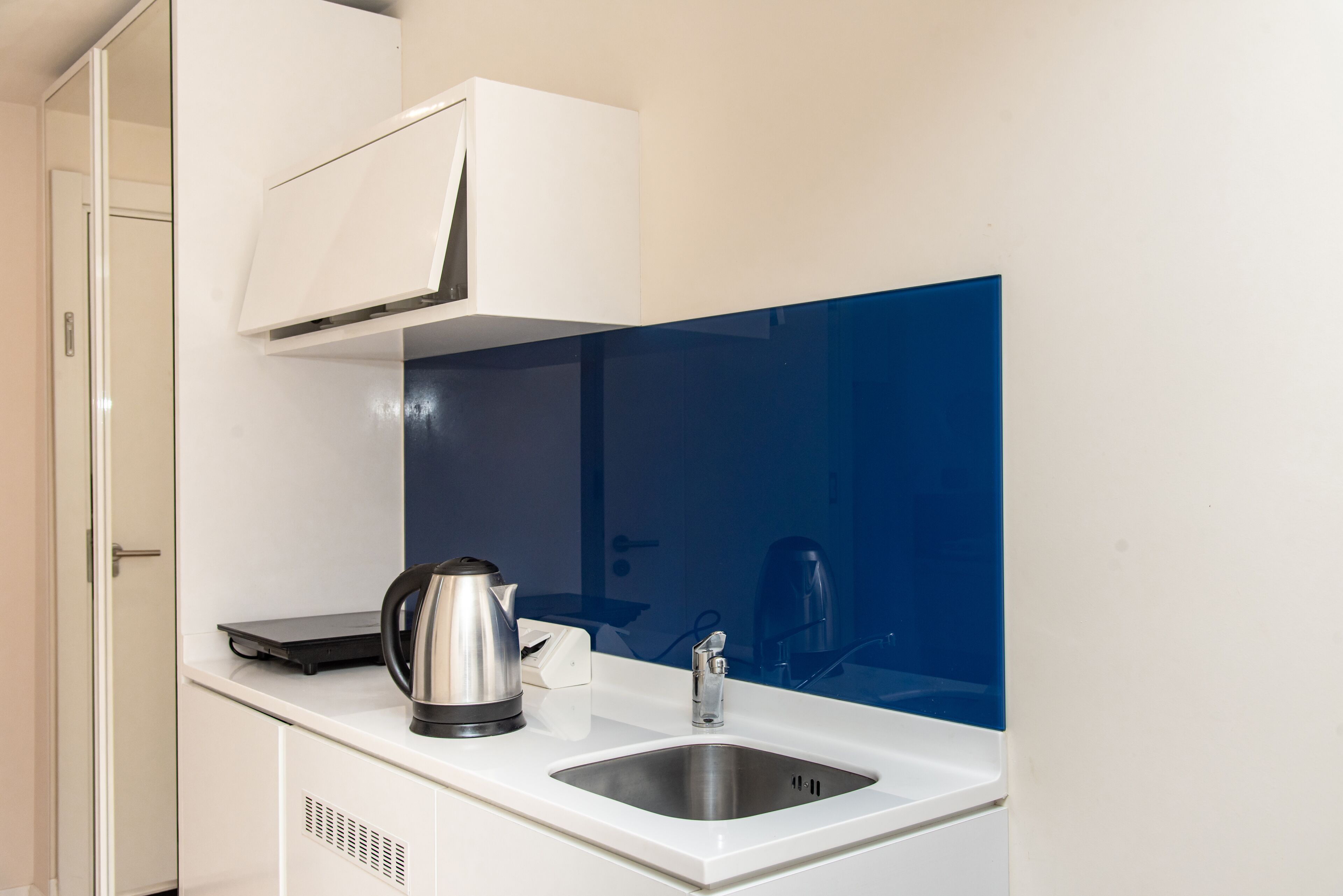 Cocina compacta con encimera blanca, pileta de acero inoxidable, tetera y cafetera.