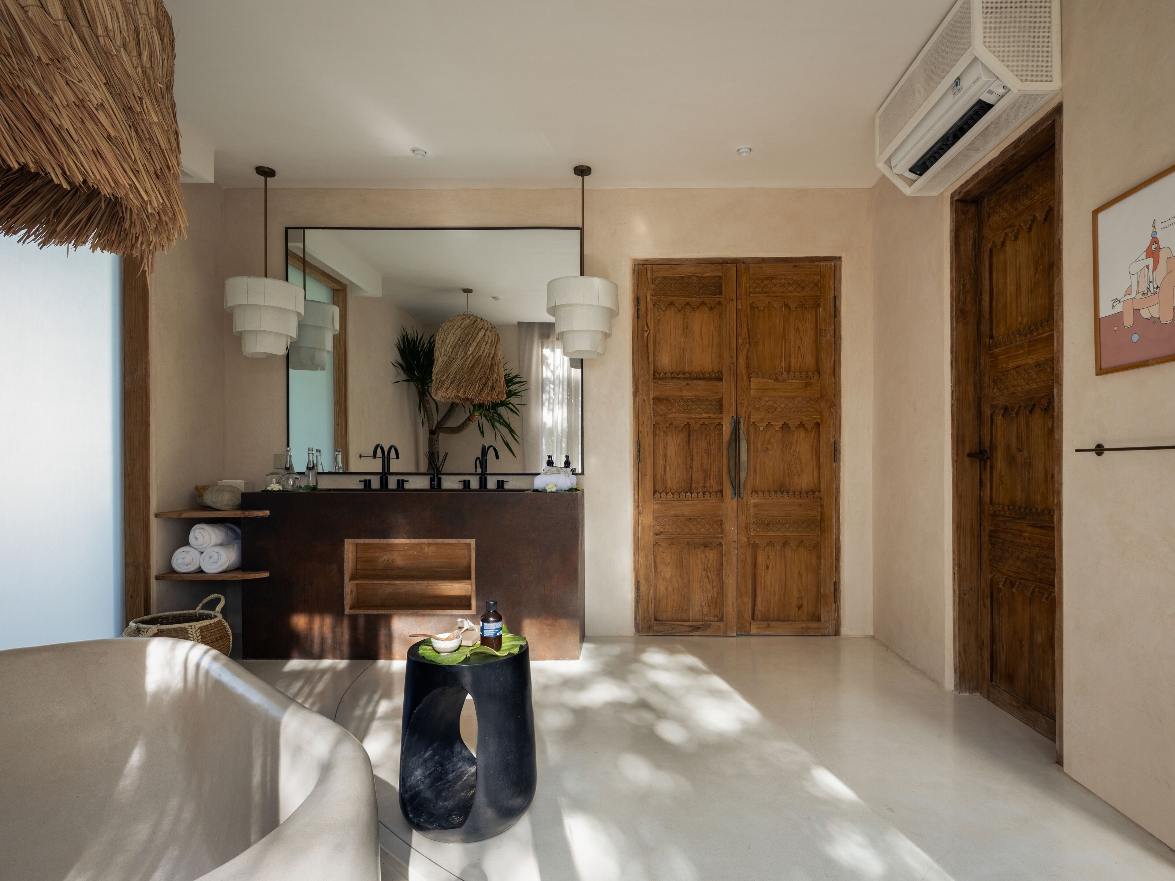 A modern bathroom with a large mirror, two sinks, a wooden door, and a black marble stool.