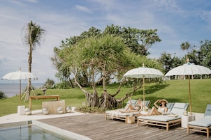 Zona de piscina con sillones reclinables, sombrillas y una terraza de madera con vistas a un paisaje cubierto de césped, árboles y el mar.