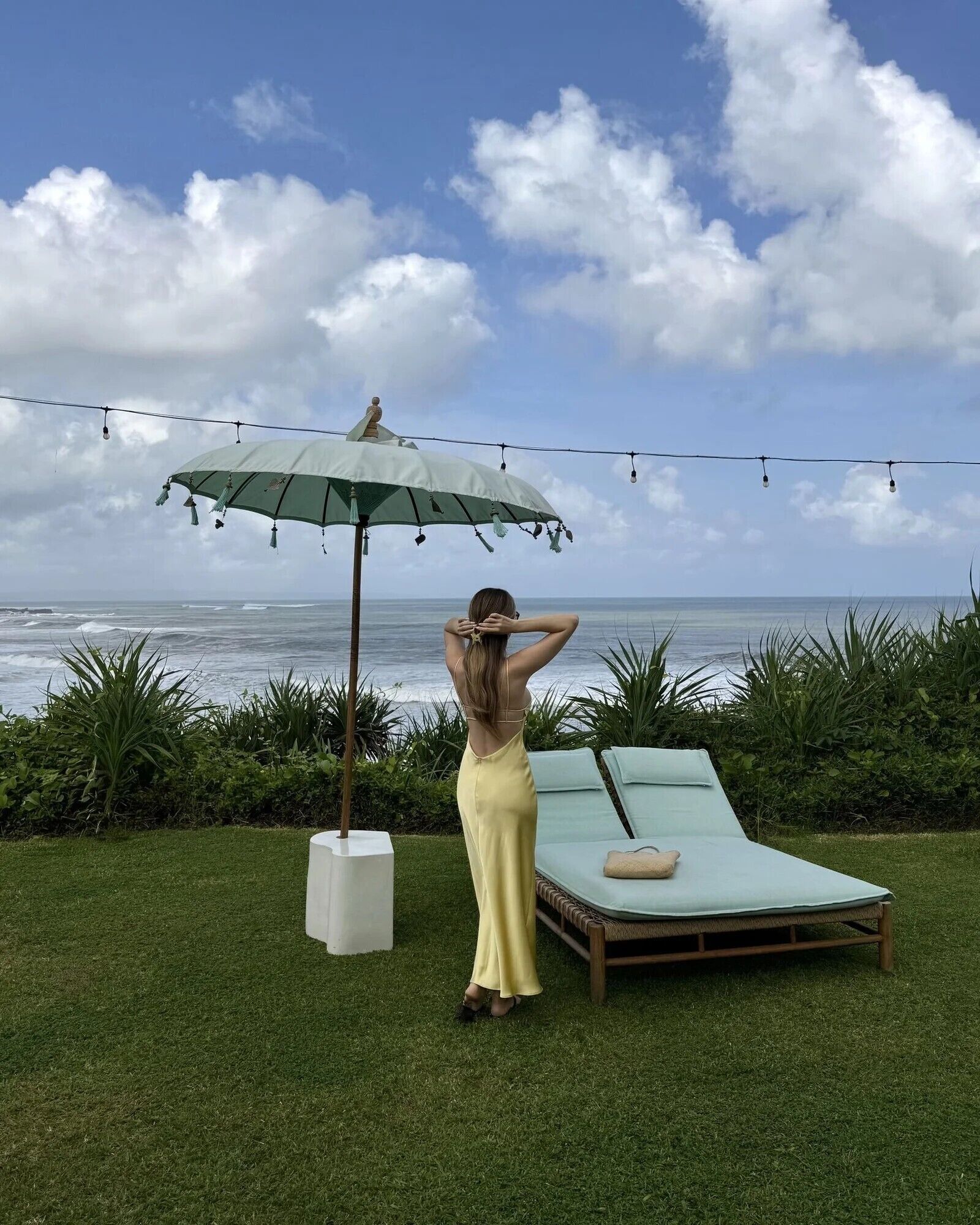 A woman standing under a green umbrella, overlooking the ocean with a beach chair and lounge chairs in the foreground.