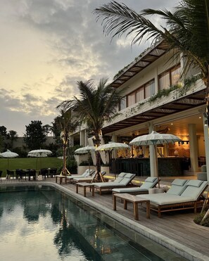 A poolside area with lounge chairs and umbrellas, a pool, and a building with a covered patio. - Oshom Bali at Nuanu Creative City (Tabanan)