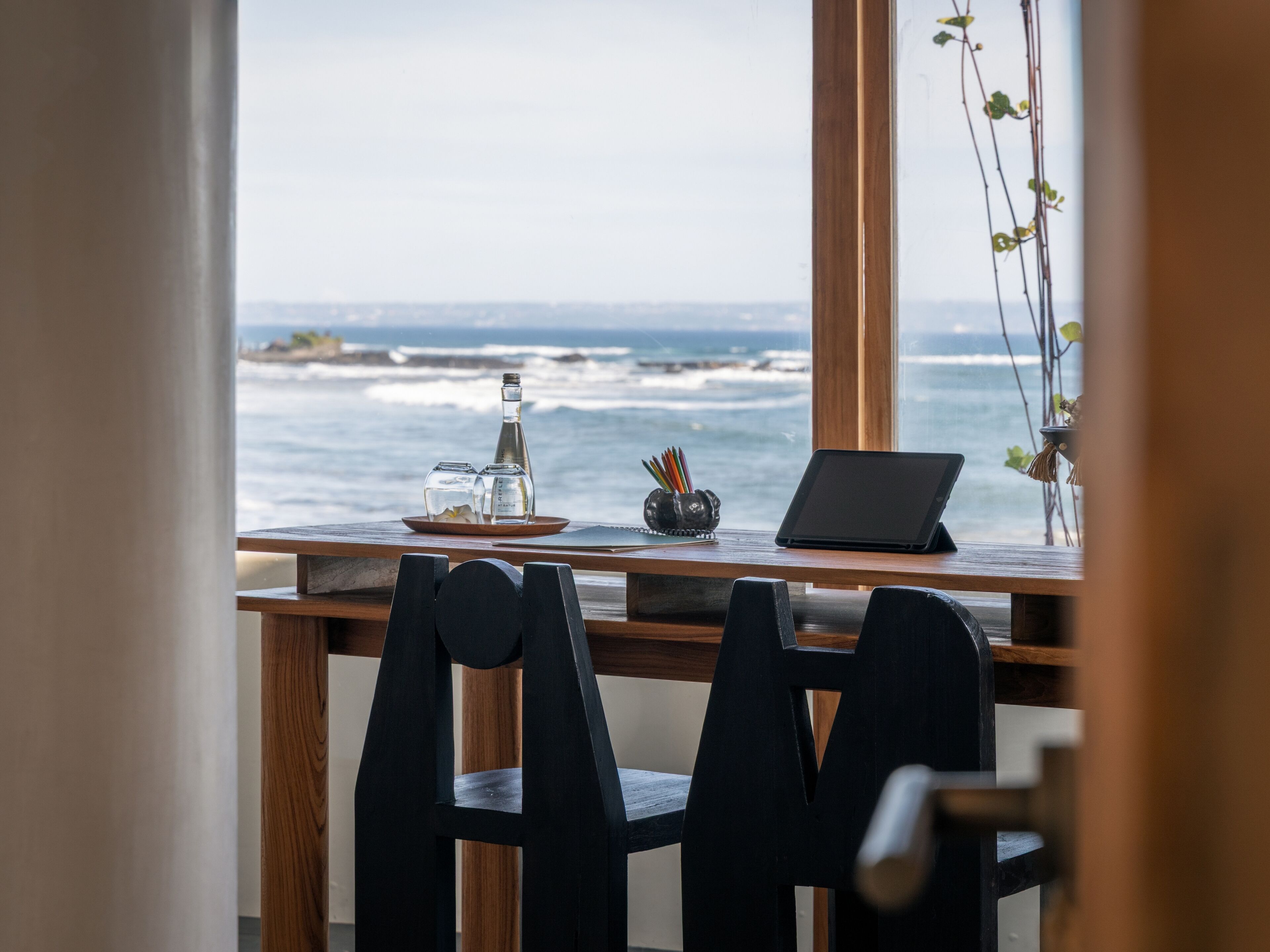 A wooden desk with a laptop, salt and pepper shakers, and a notebook by a window overlooking the ocean.