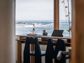 A wooden desk with a laptop, salt and pepper shakers, and a notebook by a window overlooking the ocean.