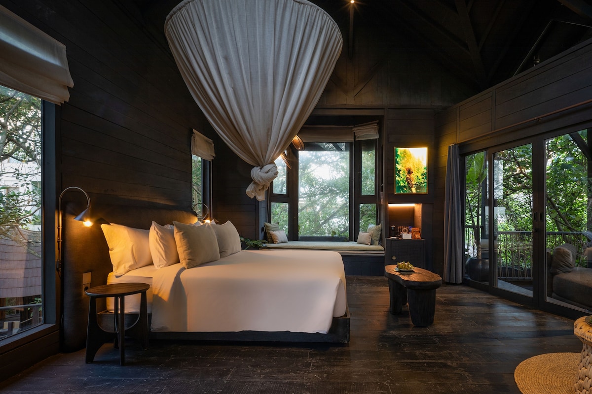 A bedroom with a large bed, a hanging light fixture, and a view of greenery outside.