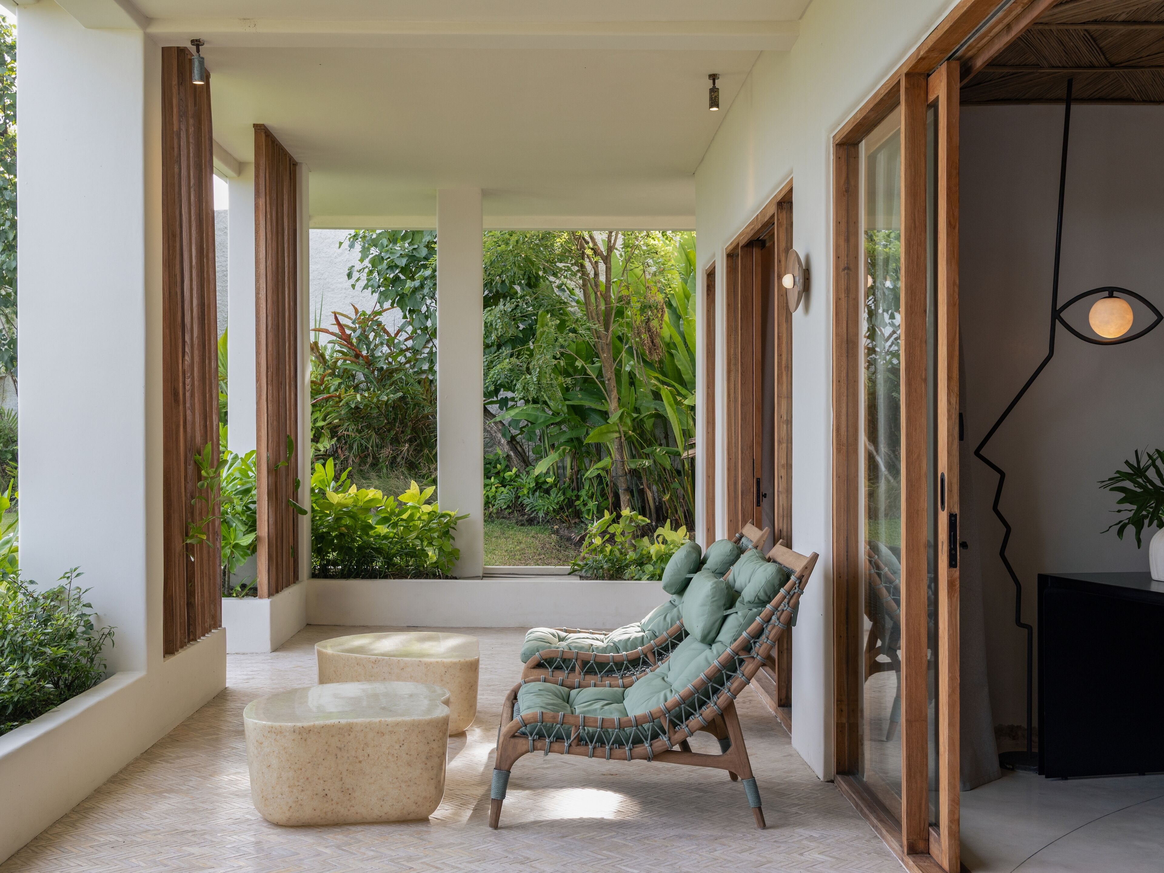 A room with a wicker chair, a round ottoman, and a glass table. There are wooden doors and a view of greenery outside.