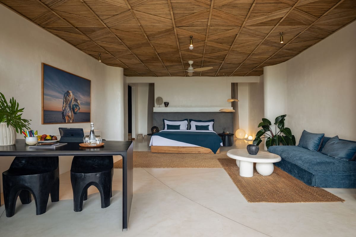 Une chambre d’hôtel moderne avec un plafond en bois, un lit avec une literie bleue, un bureau avec deux tabourets noirs, un canapé bleu et une grande plante en pot.