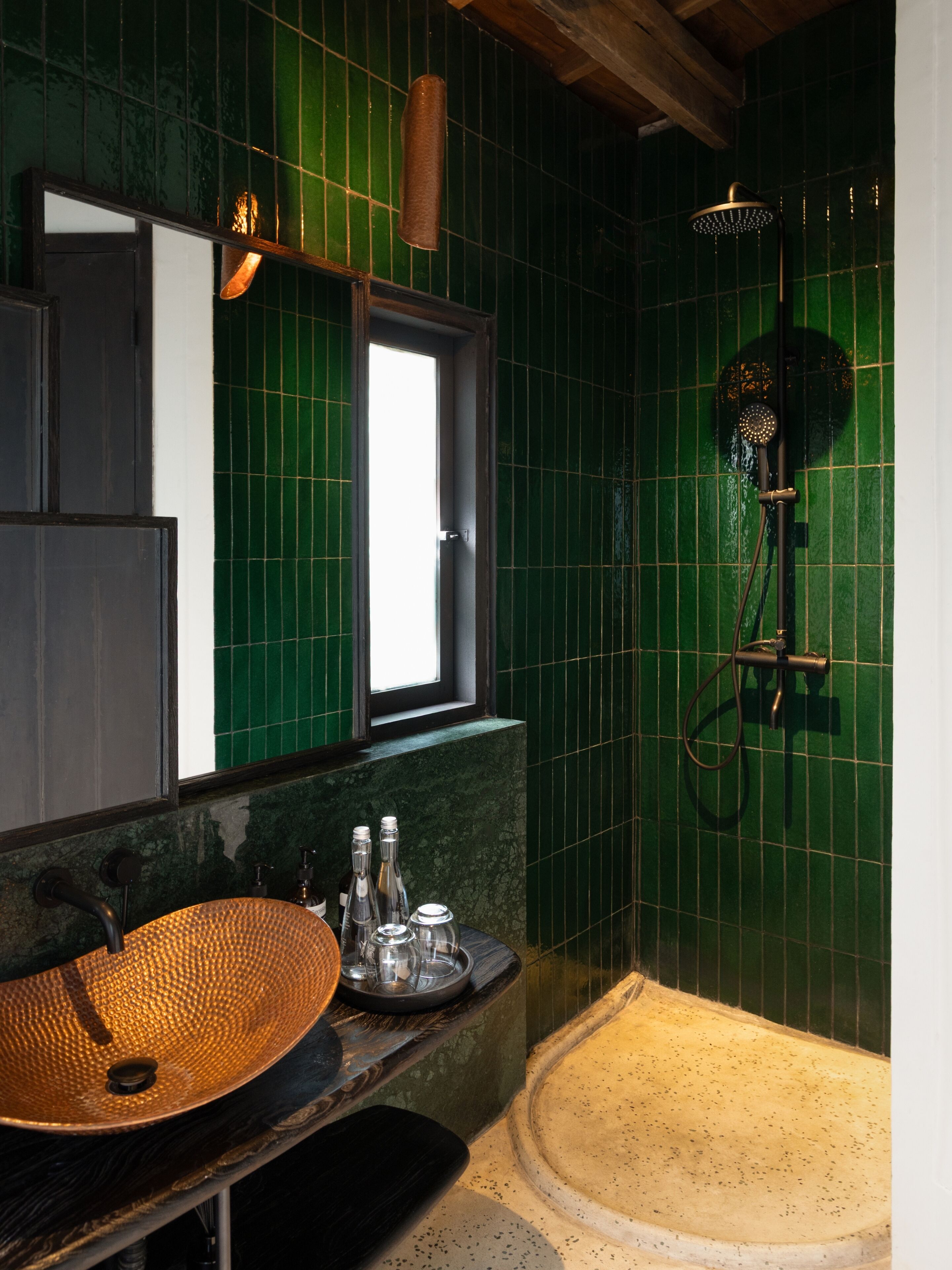 Un bagno con parete rivestita in piastrelle verdi, una panca in legno, un lavabo spazioso con una bacinella intrecciata e una doccia con soffione a mano.