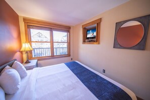 A hotel room with a bed, a TV mounted on the wall, a window with a view of a building, and a lamp on a bedside table. - Springs 8880 (Keystone)