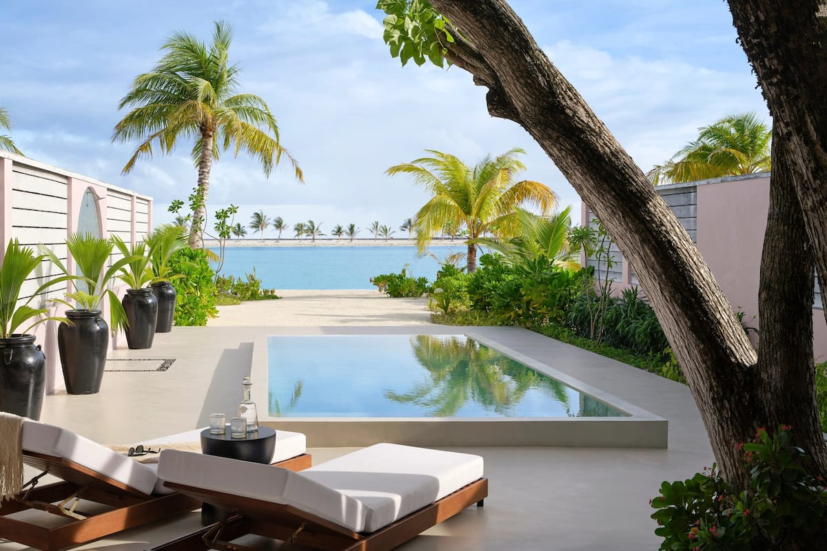 A modern poolside area with a lounge chair and a small table, surrounded by potted plants and palm trees, overlooking a beach.