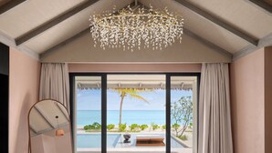 Camera da letto con un'ampia finestra che si affaccia su una piscina e una spiaggia, un letto, un lampadario sospeso e vista sull'oceano.